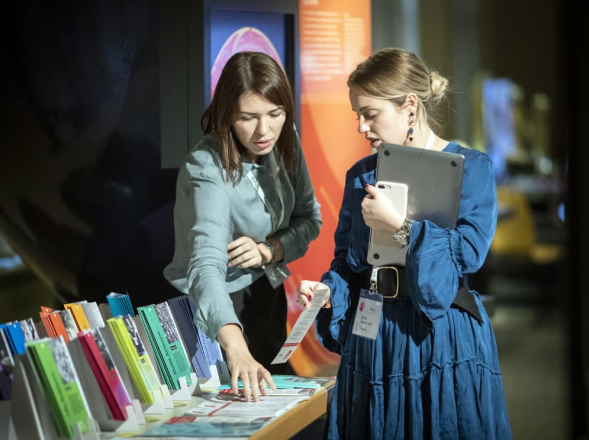 Two women are standing at a table displaying various colorful brochures. One woman is pointing at a brochure while the other holds a tablet and a brochure, looking at the table attentively. Both appear to be engaged in a discussion at Berlin Science Week.