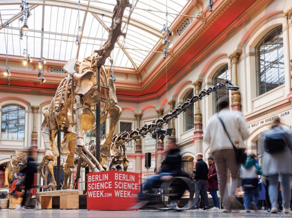 A bustling scene at Berlin's Natural History Museum during Berlin Science Week, showing blurred visitors walking past a large dinosaur skeleton exhibit, under a high arched ceiling with decorative columns. Signs promoting Berlin Science Week are visible.