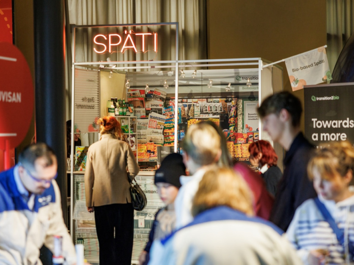 People are gathered around a small store with the sign "SPÄTI" lit in red neon above it. Displayed items, including colorful snacks, are visible inside. A person wearing a beige coat and red hair stands at the entrance, while others browse nearby kiosks or stands.
