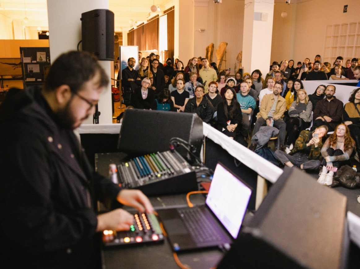A person operates audio equipment on a table with laptops and sound mixers in a dimly lit room. A large audience sits and stands attentively in the background, watching the performance. The room has a casual, informal atmosphere with various decorations and lighting.