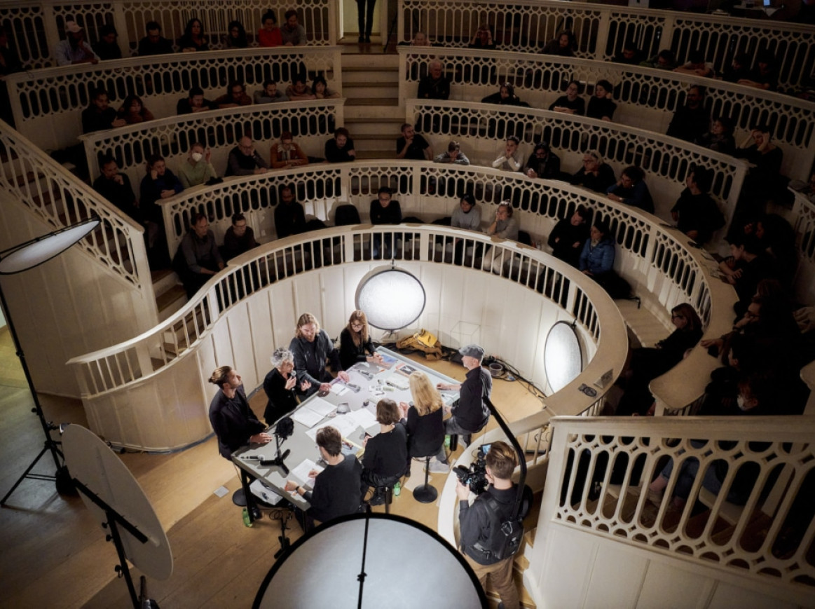 A group of people sits around a table in a circular, multi-tiered amphitheater, engaged in a discussion. The tiers are filled with spectators watching the conversation. Lighting equipment is visible around the table.