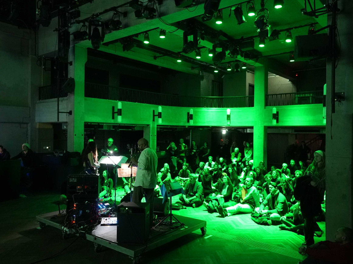 A green lit event room, with people sitting on the floor and watching a performance