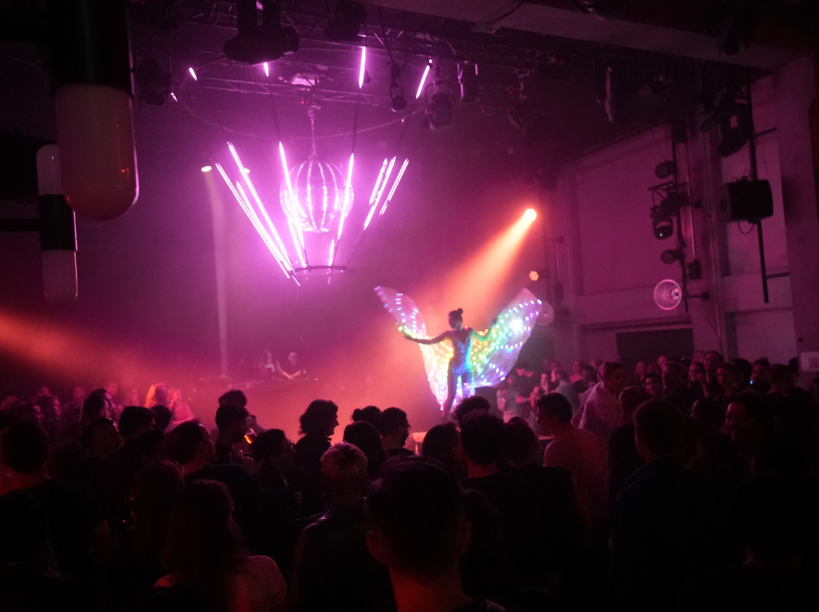 A full event room with a stage on which a person is dancing and. a light installation in the background