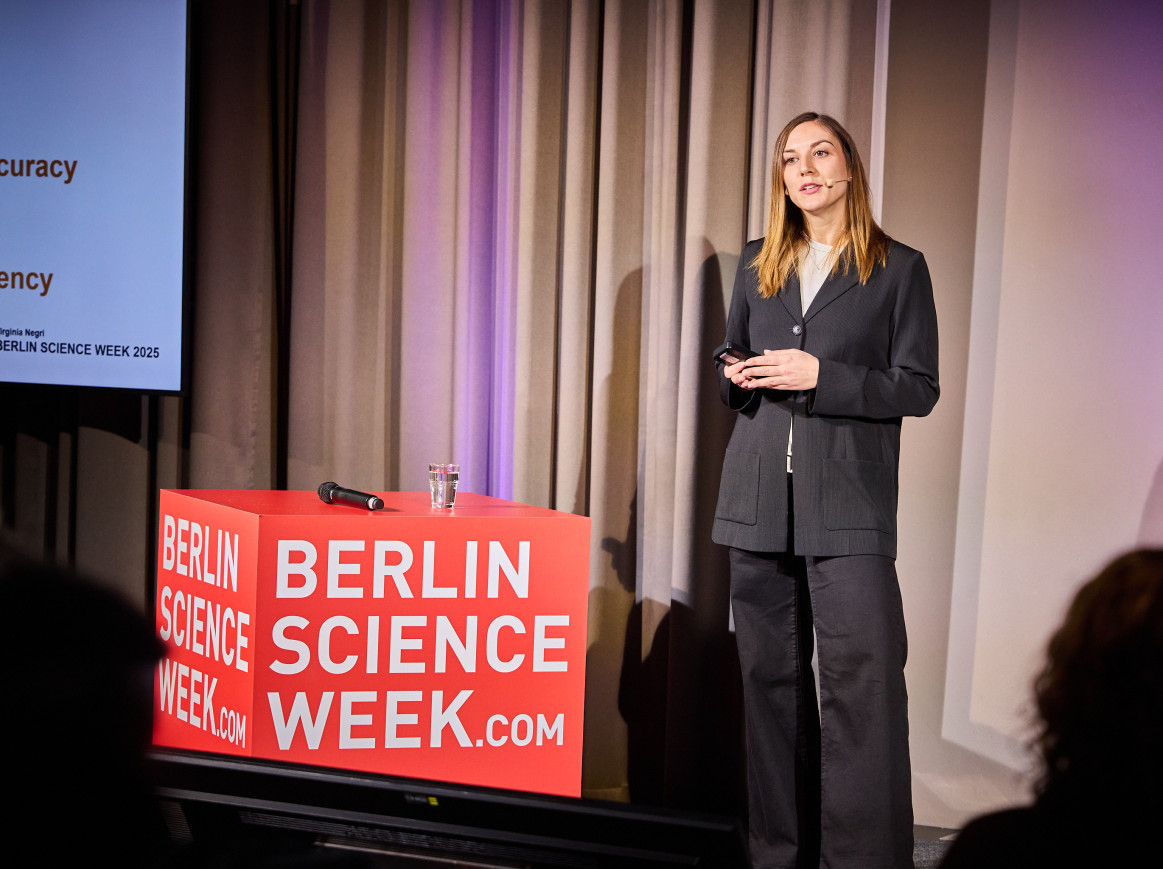 A woman stands speaking next to a big red cube. 