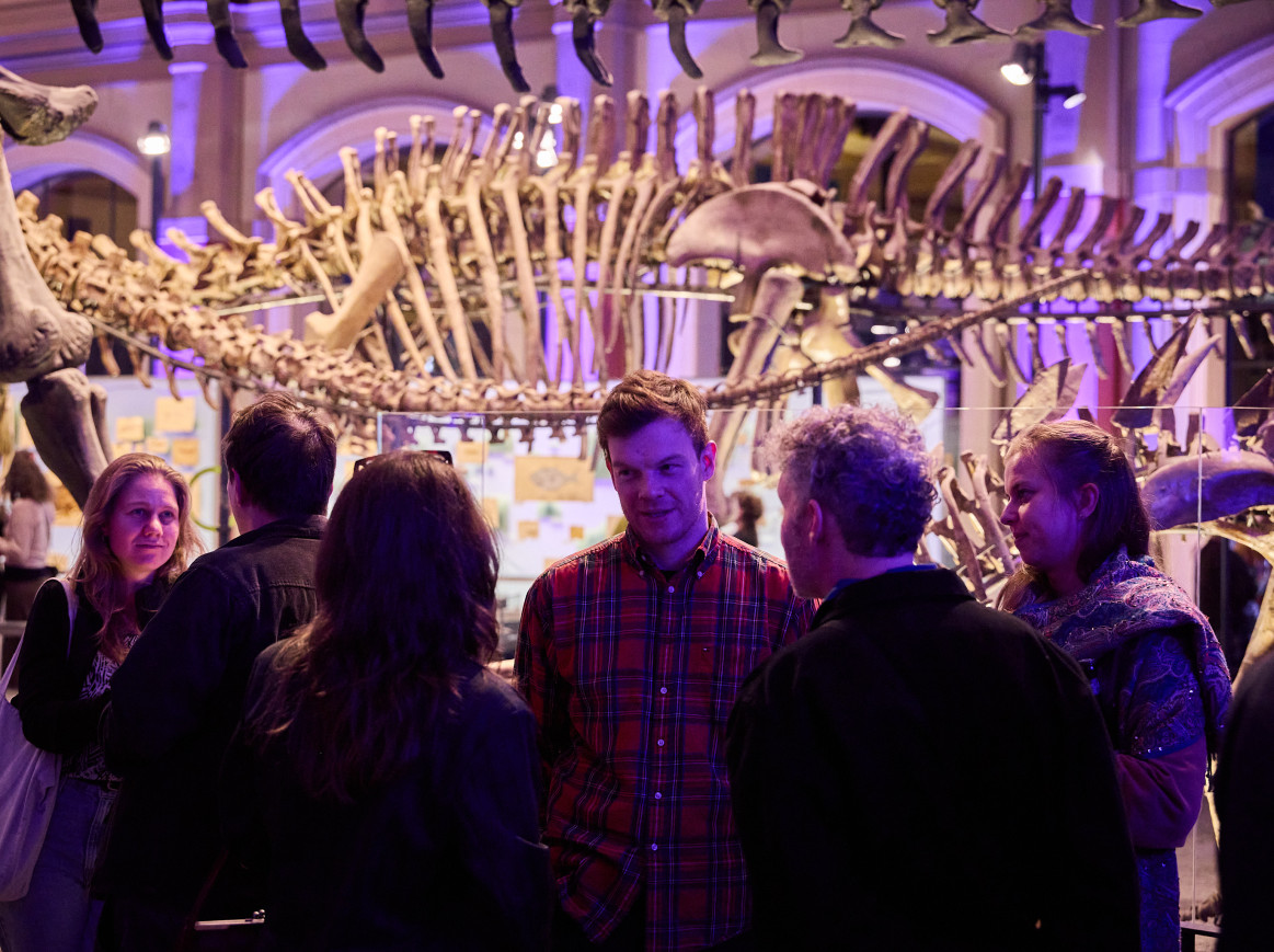 A man looks at 5 other people as he speaks to them in front of a big dinosaur skeleton. 