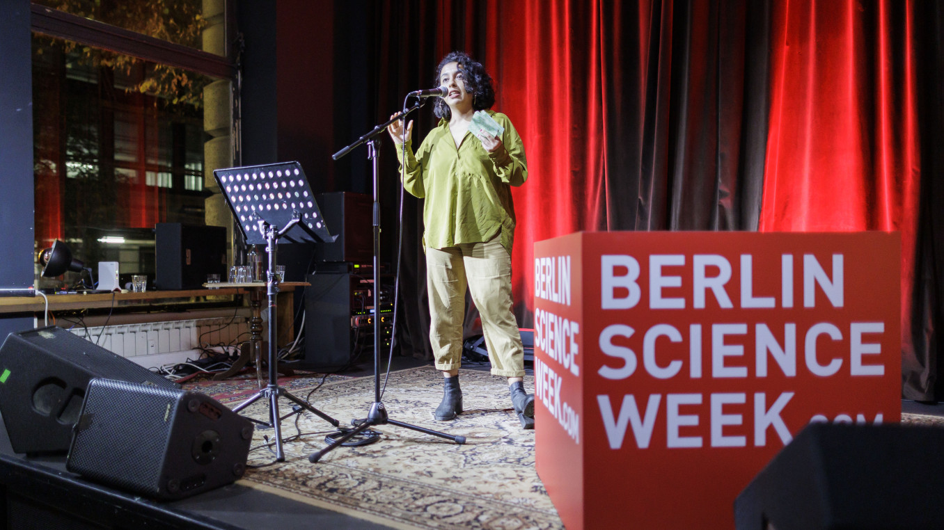 Person on stage speaking into a microphone next to a "Berlin Science Cube" cube