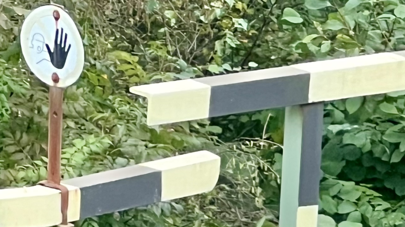 The image shows a barrier gate, likely at a road or railway crossing, painted in alternating black and white stripes. Next to the gate is a circular sign with a handprint design, suggesting a stop or caution signal. The background is filled with dense green foliage, indicating a natural or rural setting. The design of the gate and the sign imply a request to halt, possibly to indicate restricted access or a need for caution at the crossing.