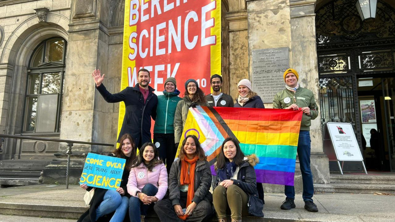 Soapbox Science Berlin and LGBTQ+ STEM Berlin teams pose with pride flag in front of Museum für Naturkunde.