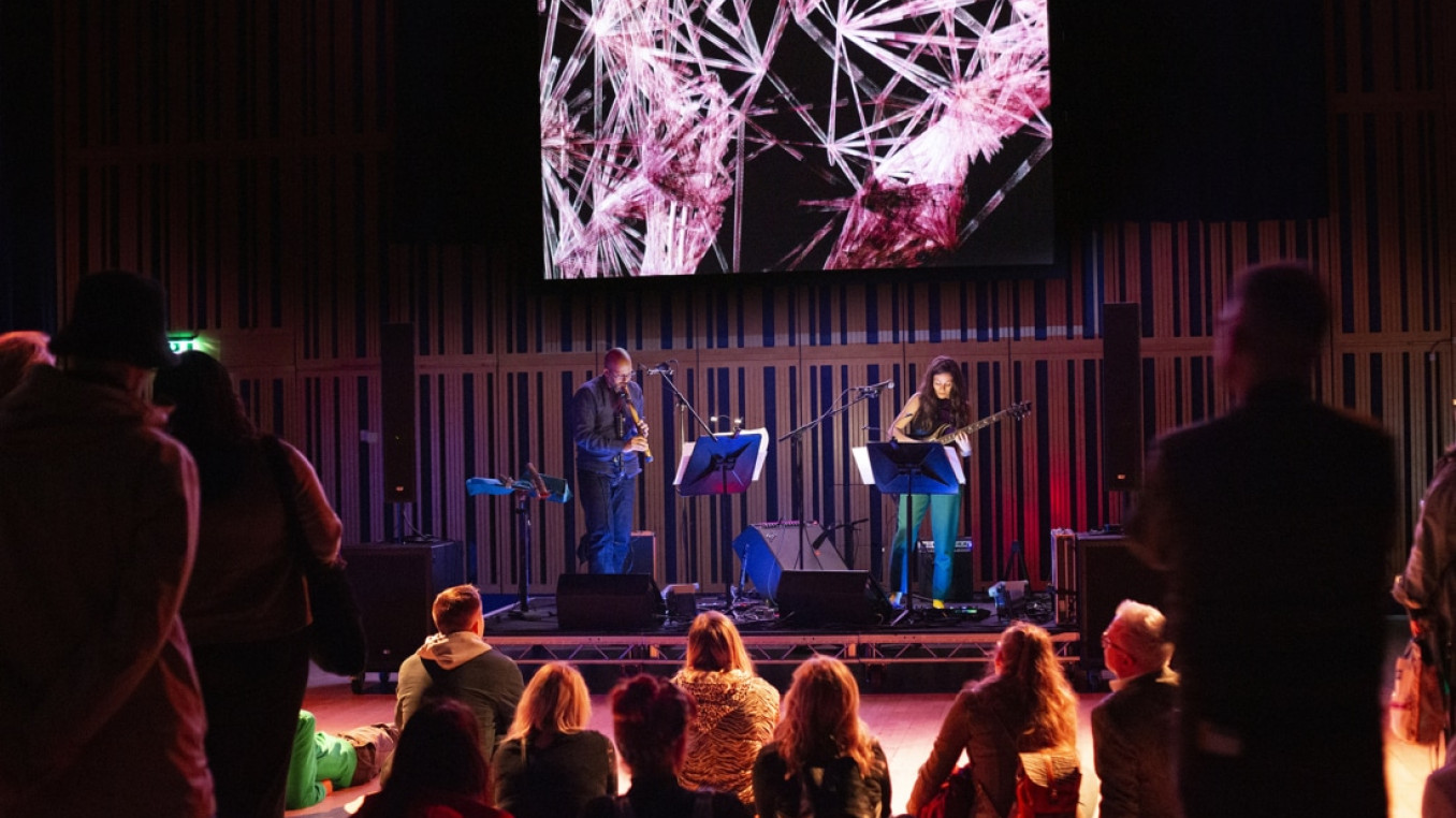 Two people on a stage playing instruments in front of a small crowd.
