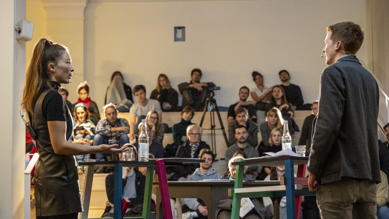 Two people facing one another stand in front of an audience. 