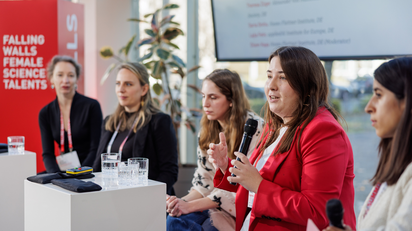 A group of five women scientists are sitting in a row. The second woman from the right is speaking. Everyone is listening to her.