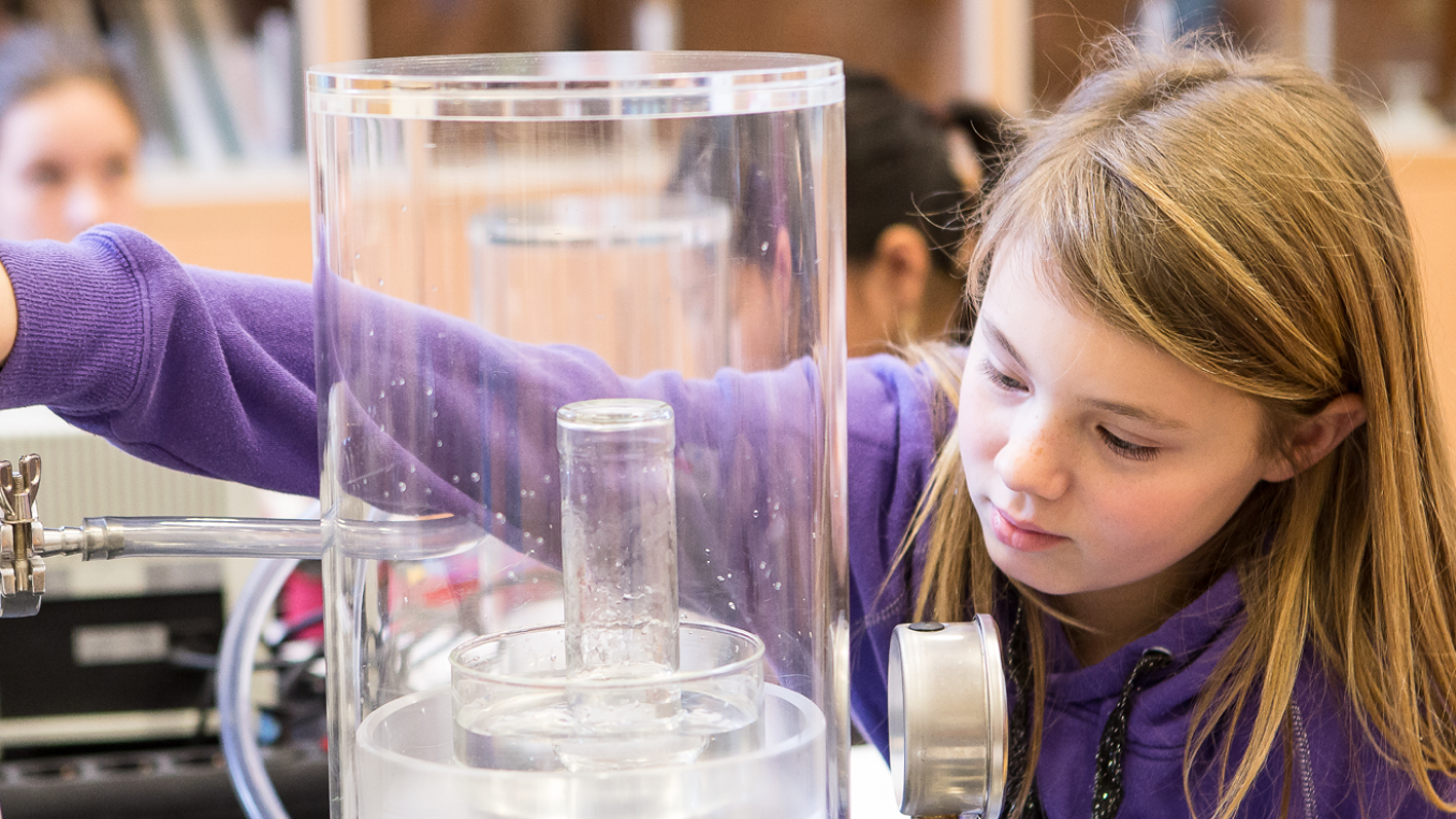 This image shows a young girl conducting a physics experiment with a vacuum bell jar at a laboratory table. It is a vacuum experiment in which the air is pumped out of a container.
