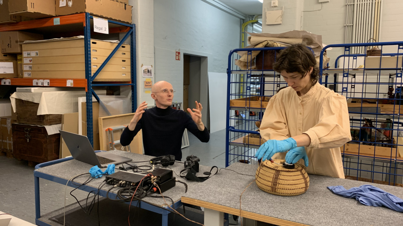 Two people are in a museum storage. One on the left side is seated and talking. One on the righ side standing and using a sound device over a museum collection item.