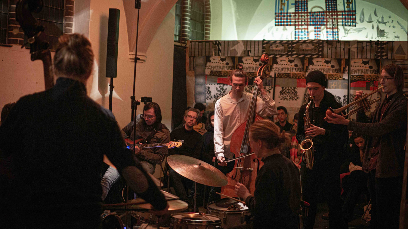 Eine kleine Jazzband spielt in einem gemütlichen, stimmungsvoll beleuchteten Raum vor einem aufmerksam lauschenden Publikum.