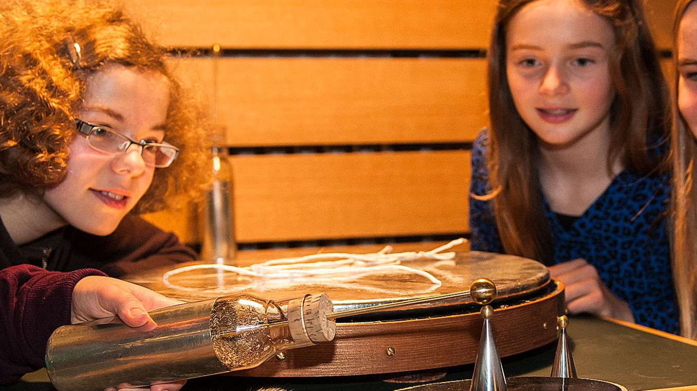 Fünf Mädchen experimentieren an einem Tisch. Ein Mädchen hält eine Glasflasche, aus der ein Stab an der Spitze ragt, an einen kleinen Metallkegel, der auf einer kleinen runden Platte mit glatter, schwarzer Oberfläche steht.