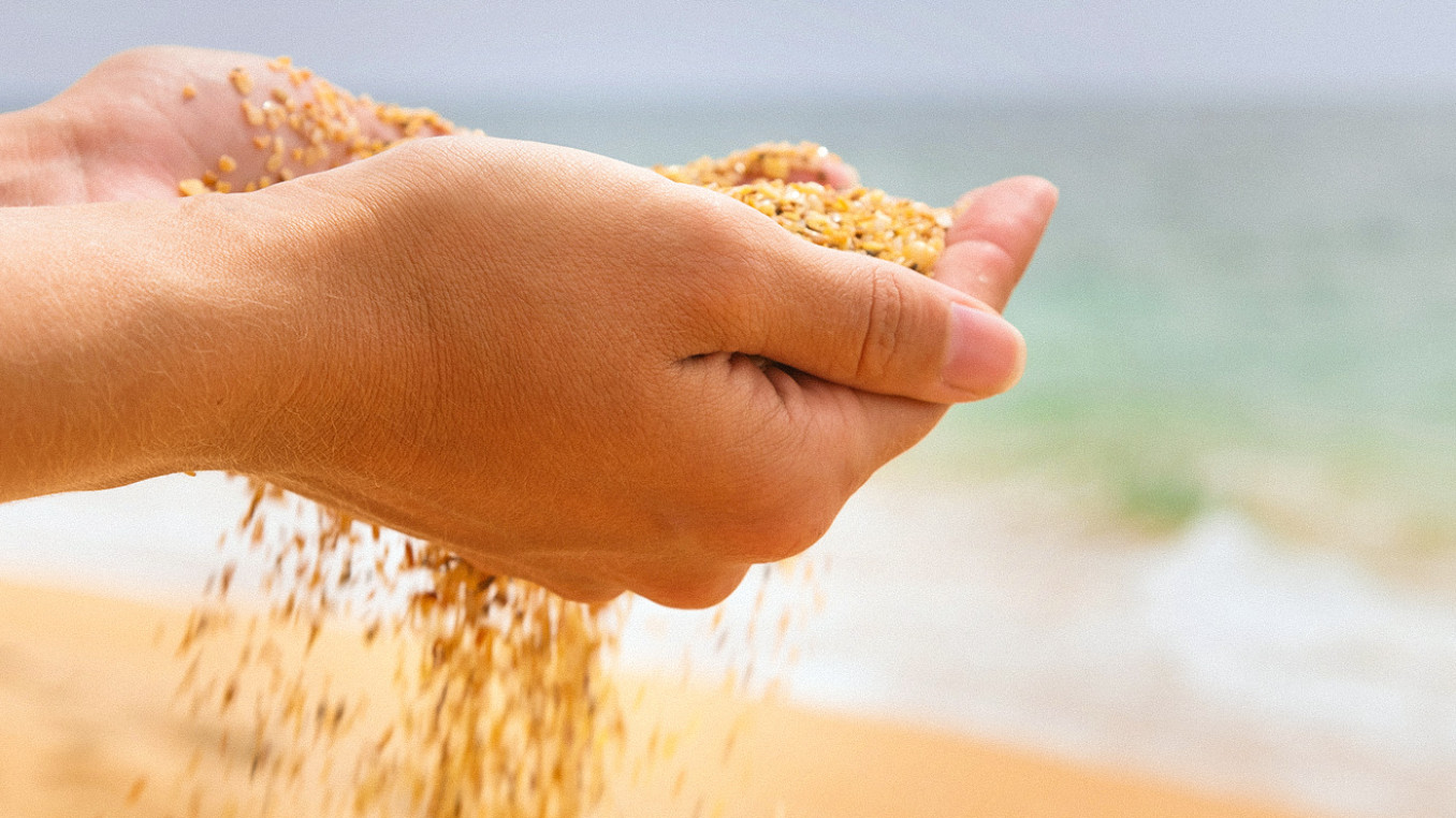 Zwei Hände halten eine Handvoll grobkörnigen Sand am Strand, während dieser langsam zwischen den Fingern hindurchrieselt. Im unscharfen Hintergrund sind das Meer und die sandige Küstenlinie bei hellem Tageslicht zu erkennen.