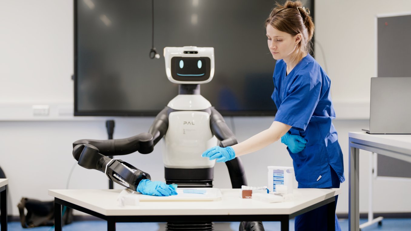 Der humanoide Roboter TIAGo Pro steht an einem Arbeitstisch mit Tabletten-Sortierschachteln, eine Person greift ebenfalls auf den Tisch zu.