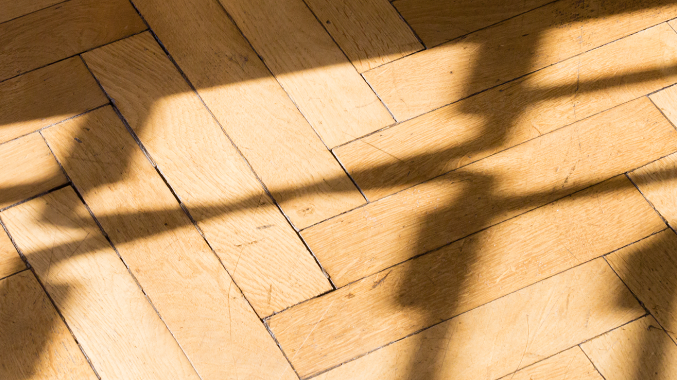Nahaufnahme eines Parkettbodens aus hellem Holz im Fischgrätmuster. Über die Dielen fallen scharf gezeichnete Schatten einer schmiedeeisernen Balustrade, die durch Sonnenlicht abstrakte, geschwungene Linienmuster auf das Holz wirft.