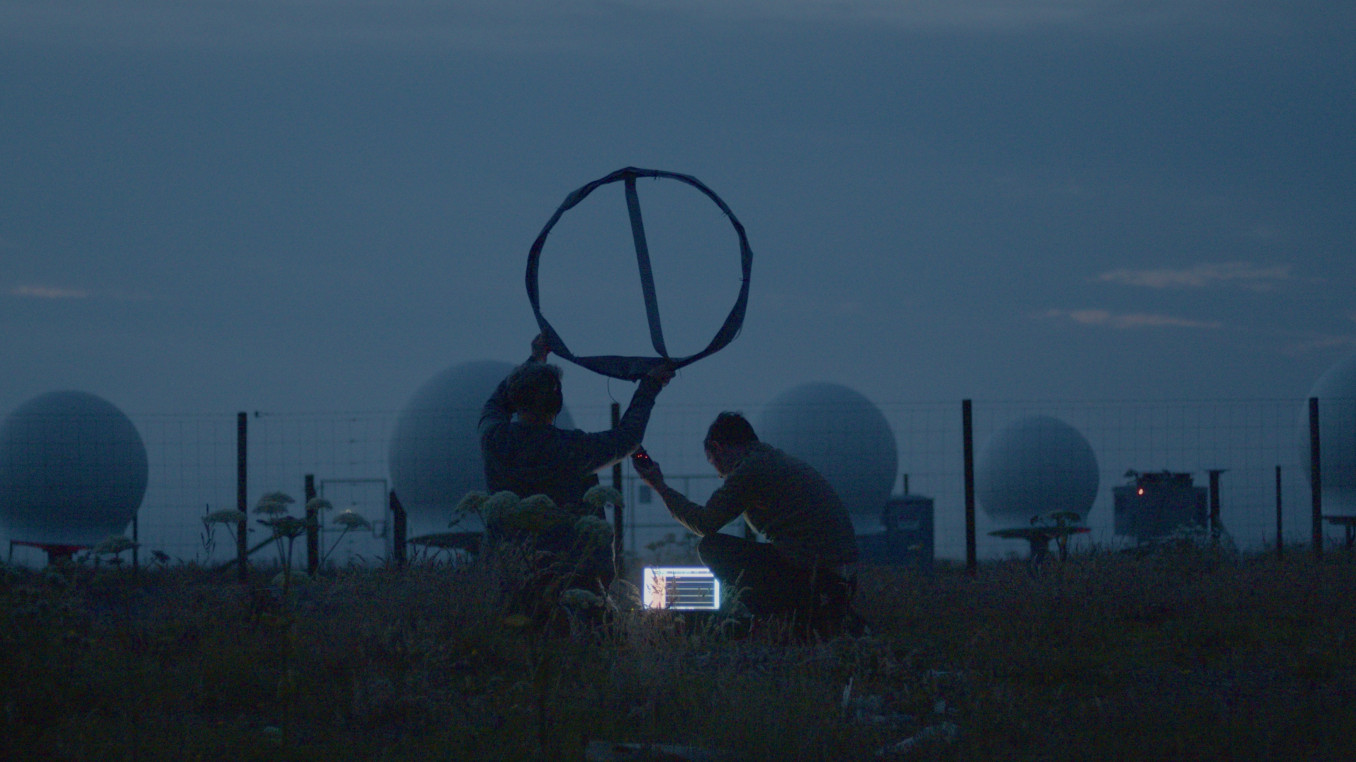 Image from Song for the Cosmos showing artist Edwin Mingard and musician Gina Ritch at dusk, listening to a spaceport's ground control using an experimental aerial