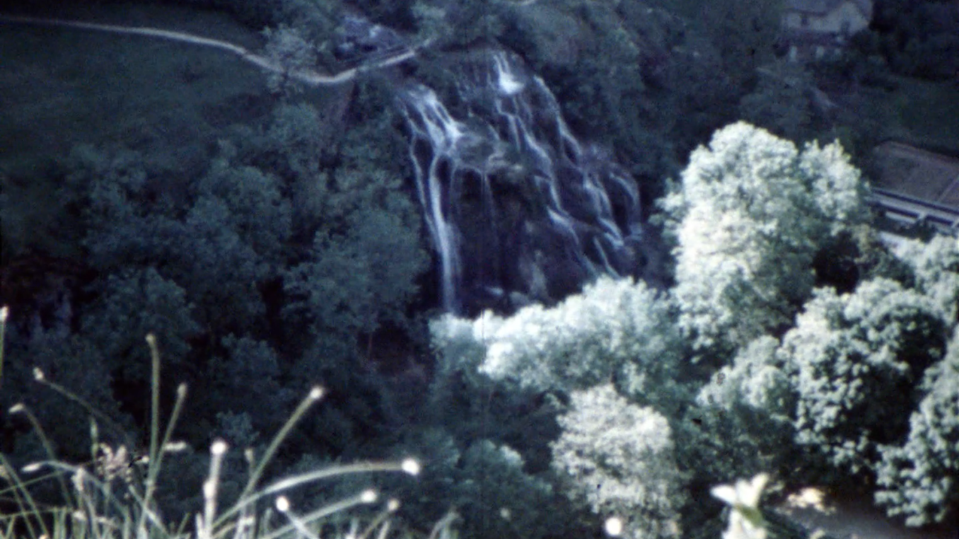 A film still from archival footage showing a forested valley, a waterfall and a house in the distance. 