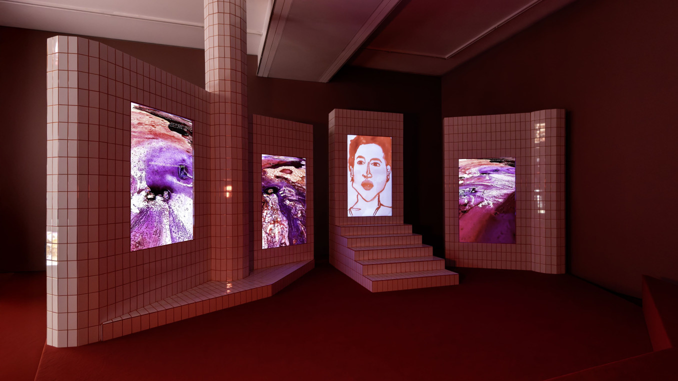 Four vertical screens in white tiles with red grout show videos of watercolor images and a portrait.